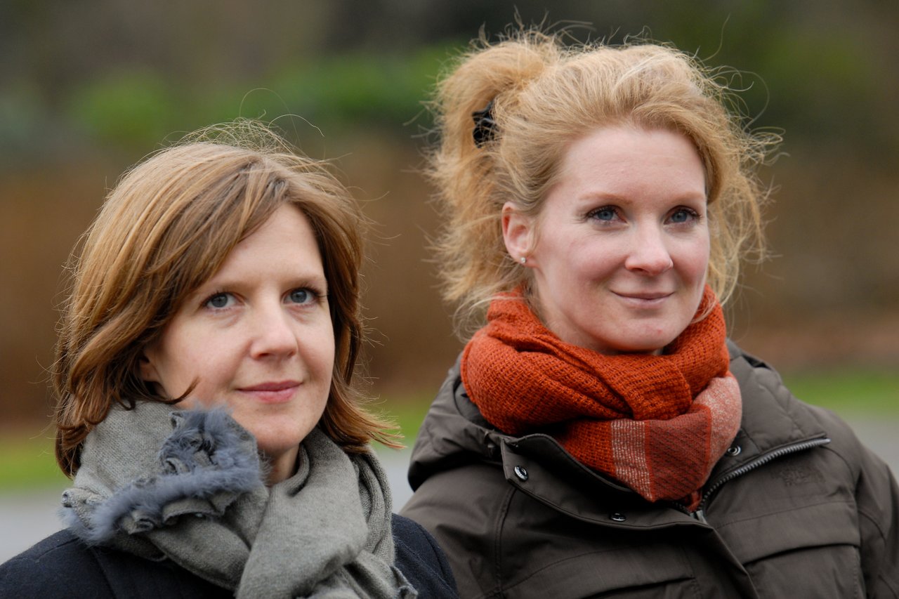 Two women wearing warm scarves and jackets stand outdoors, looking ahead with neutral expressions.