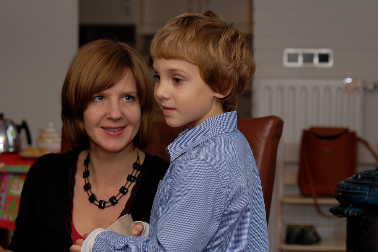 A woman smiles while sitting behind a young boy in a blue shirt, who looks off to the side.