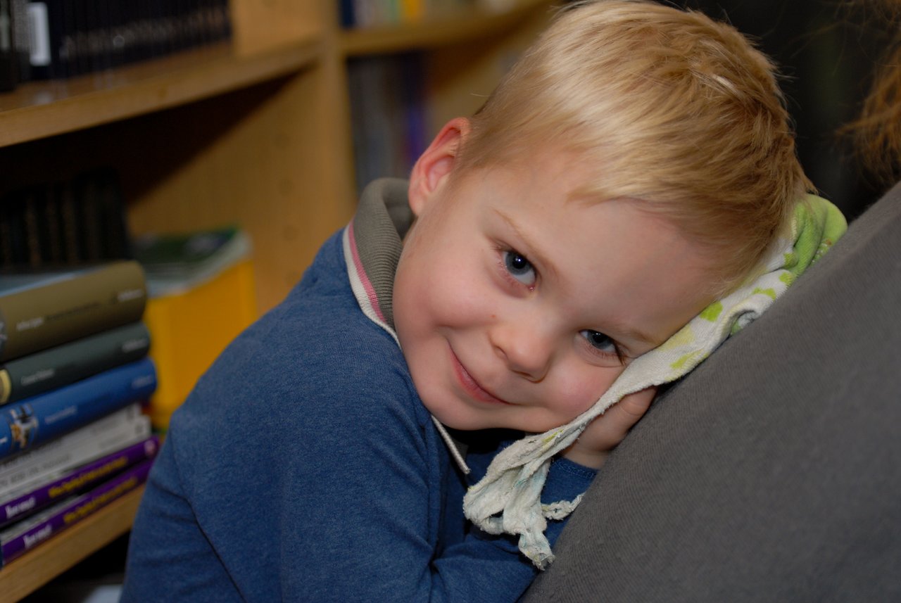 A young child with blond hair leans on someone's shoulder, resting their head on a small blanket and smiling.