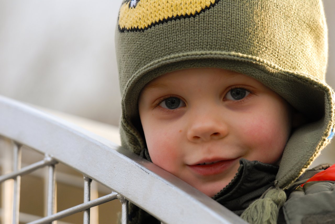 A child wearing a green winter hat smiles while leaning on a metal railing.