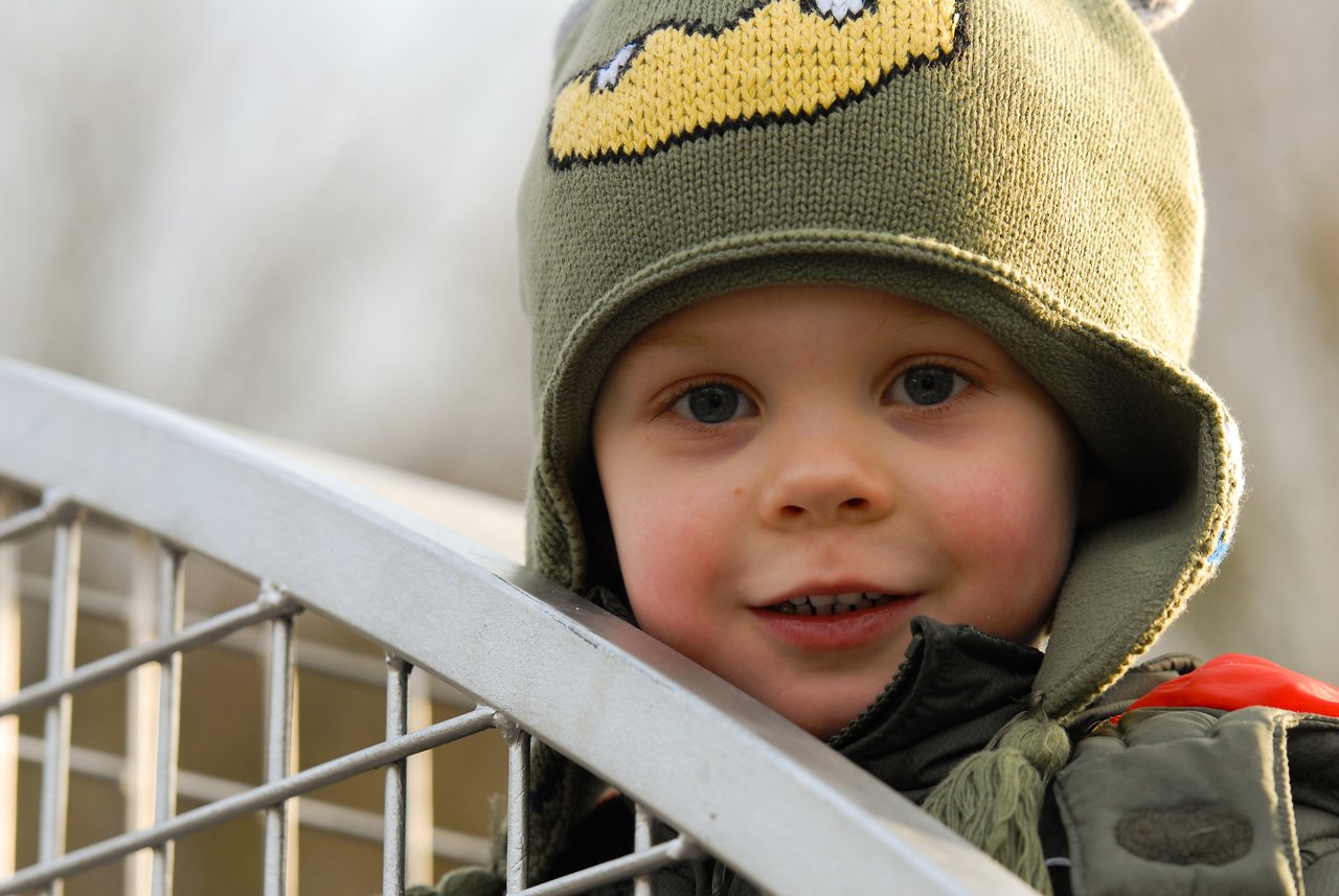 A young child wearing a green hat with a face design smiles while leaning on a metal railing outdoors.