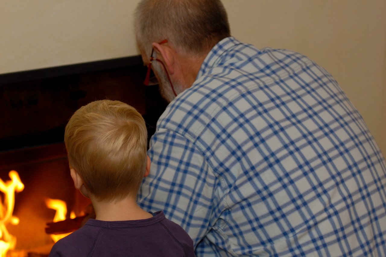 A child and an older man sit close to a fireplace, watching the flames together.