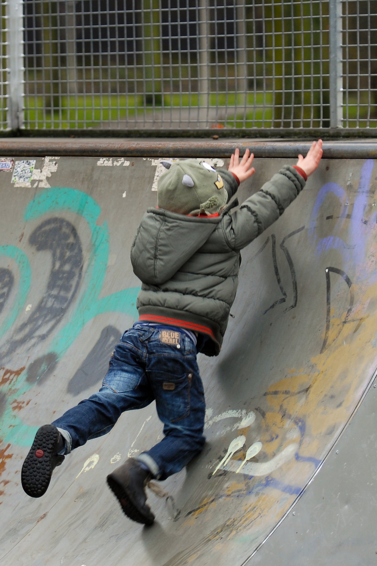 A child in a green jacket and hat climbs a graffiti-covered half-pipe, reaching for the top edge.