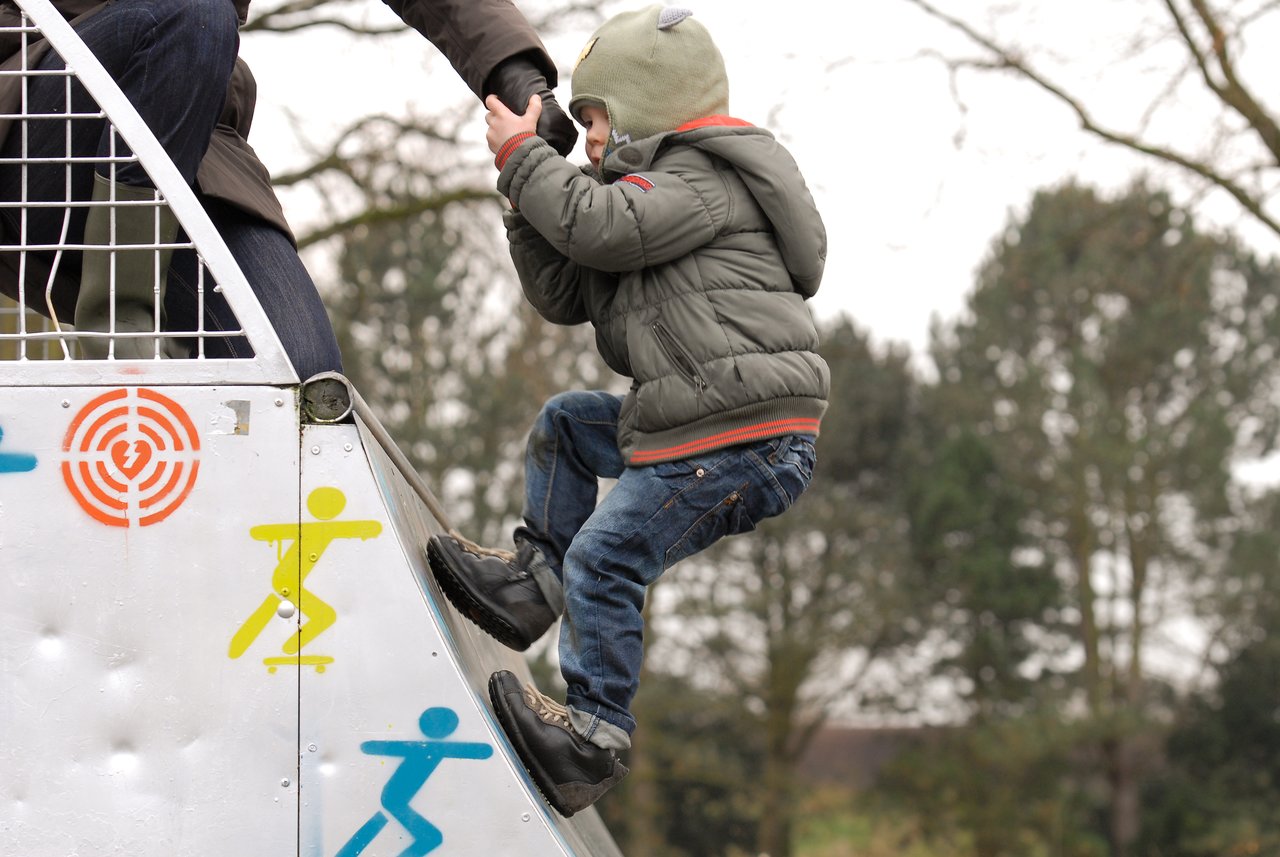 A child in a green jacket climbs a half-pipe, holding an adult's hand for support.