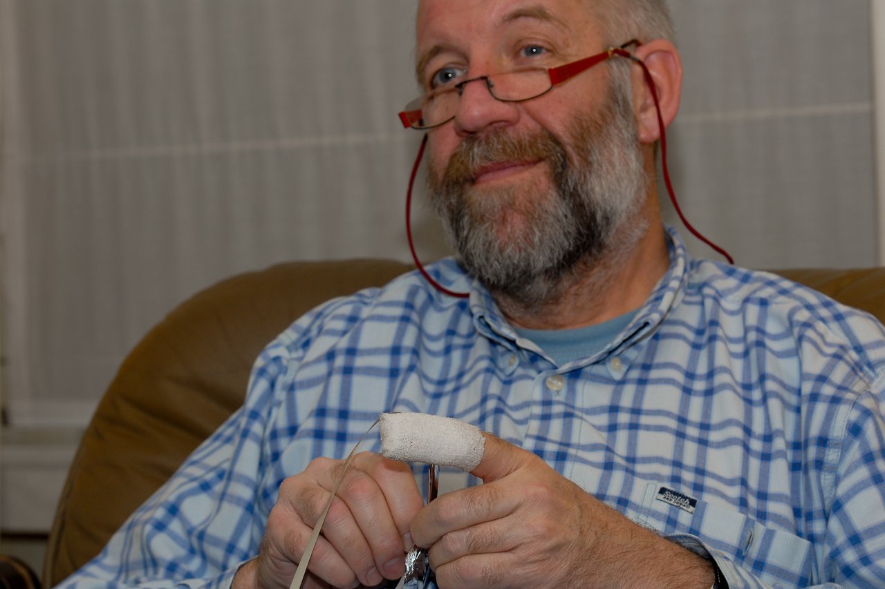 A man with glasses and a beard sits on a couch, holding an object with a bandaged finger.