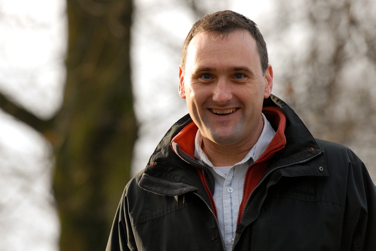 A man in a black jacket smiles while standing outdoors with trees in the background.