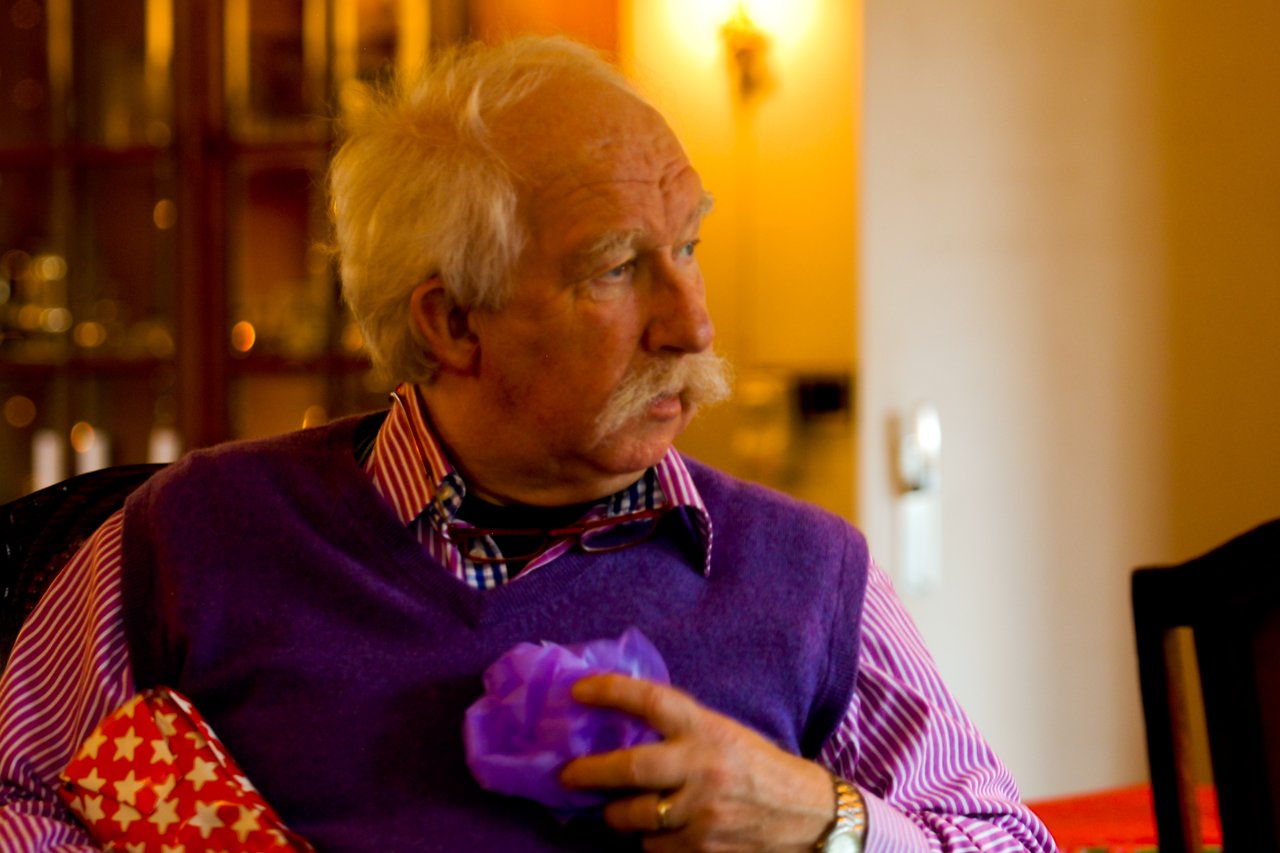 An older man in a purple sweater holds a small gift while looking to the side.