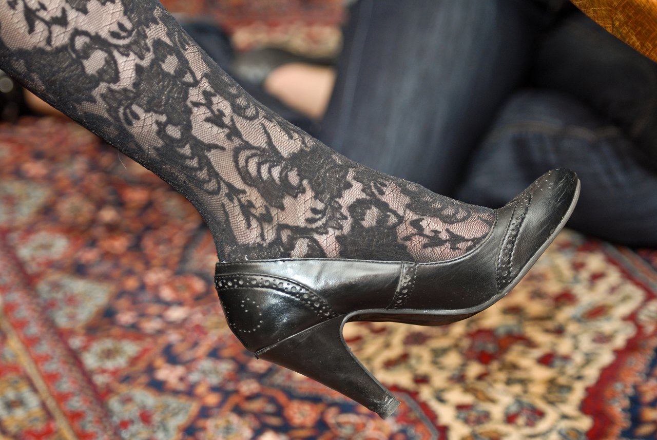 A close-up of a leg wearing black lace-patterned tights and a black high-heeled shoe on a patterned rug.