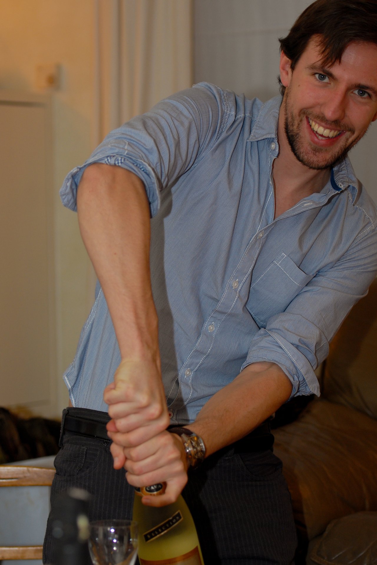 A man in a blue shirt smiles while opening a bottle of champagne with both hands.