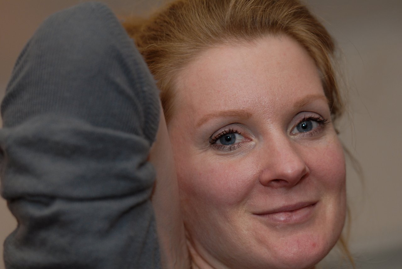 A woman with red hair smiles at the camera, with a blurred arm and sleeve in the foreground.