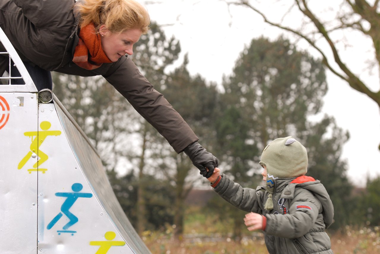 A woman leans down from a skate ramp, reaching out to hold a child's hand.