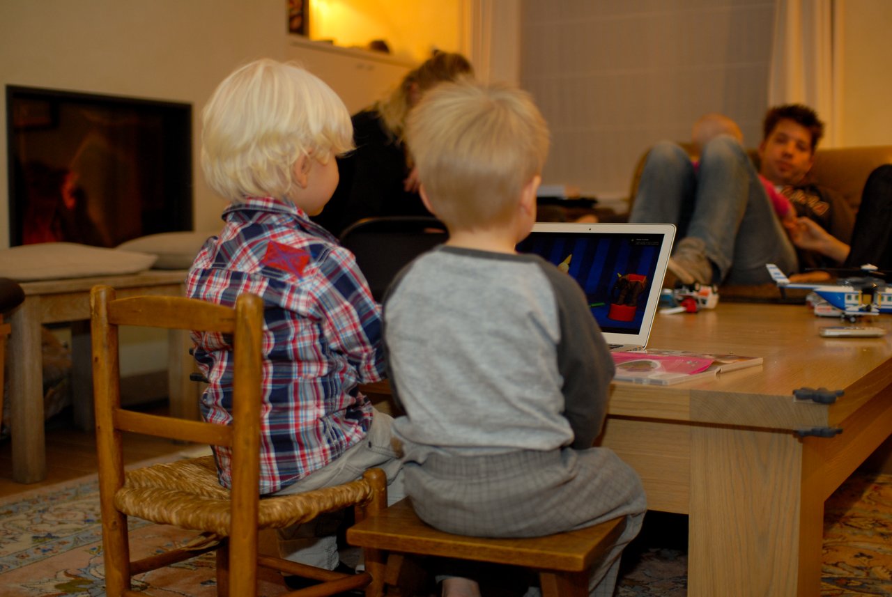 Two young children sit on small chairs watching a laptop screen, while adults relax in the background.