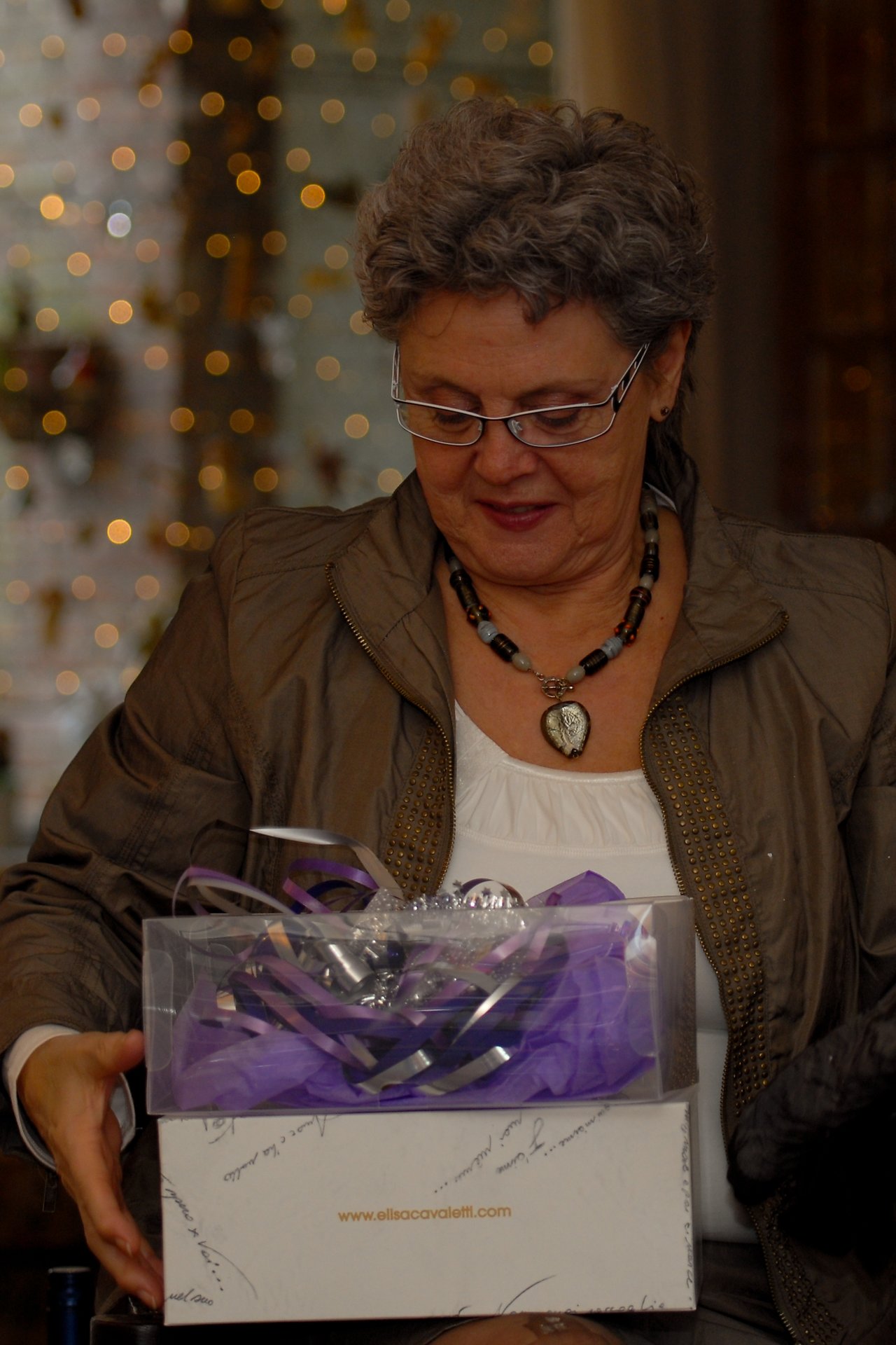 A woman wearing glasses and a necklace looks down at a decorated gift box in her hands.