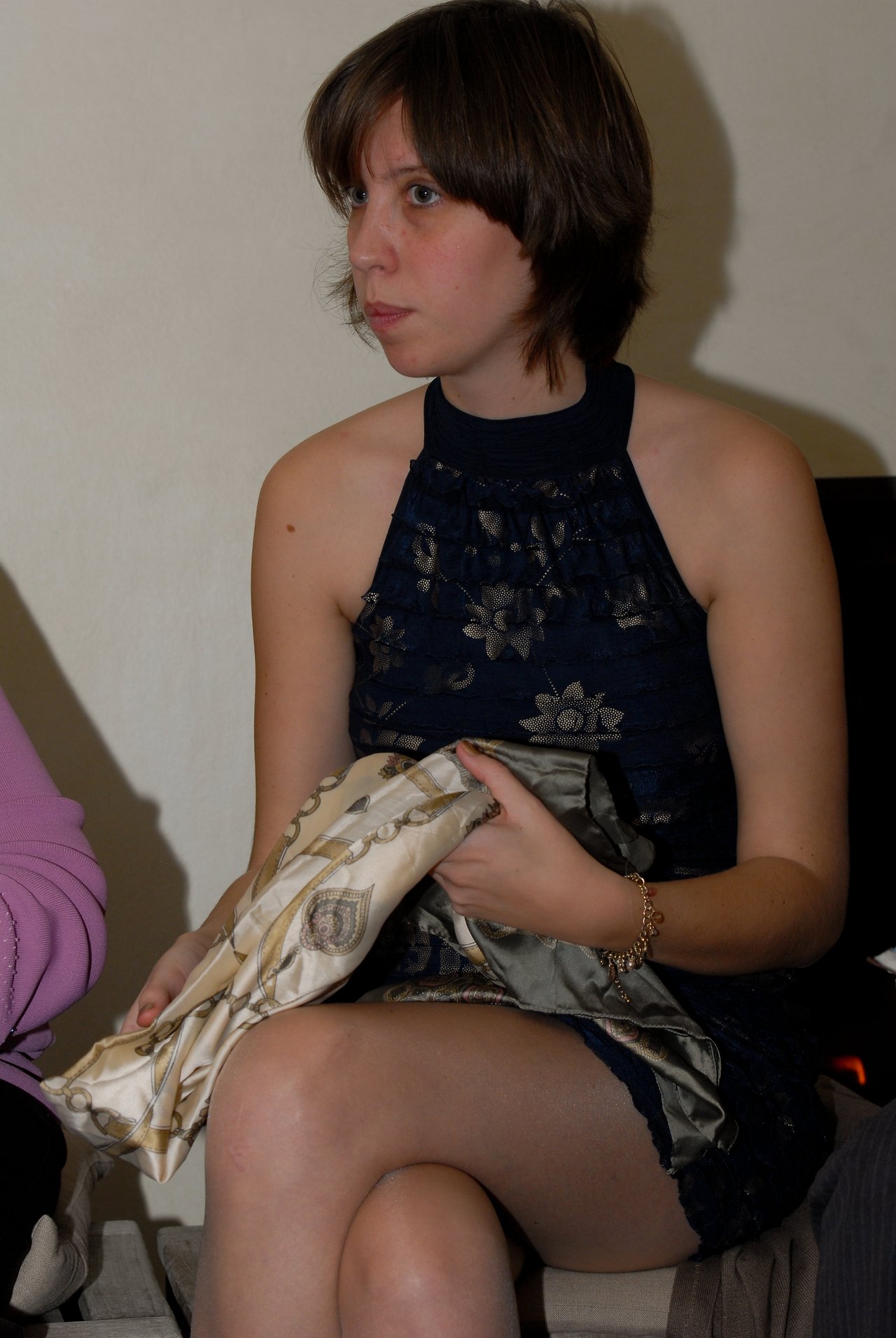 A woman in a dark blue dress sits holding a patterned fabric, looking to the side with a neutral expression.