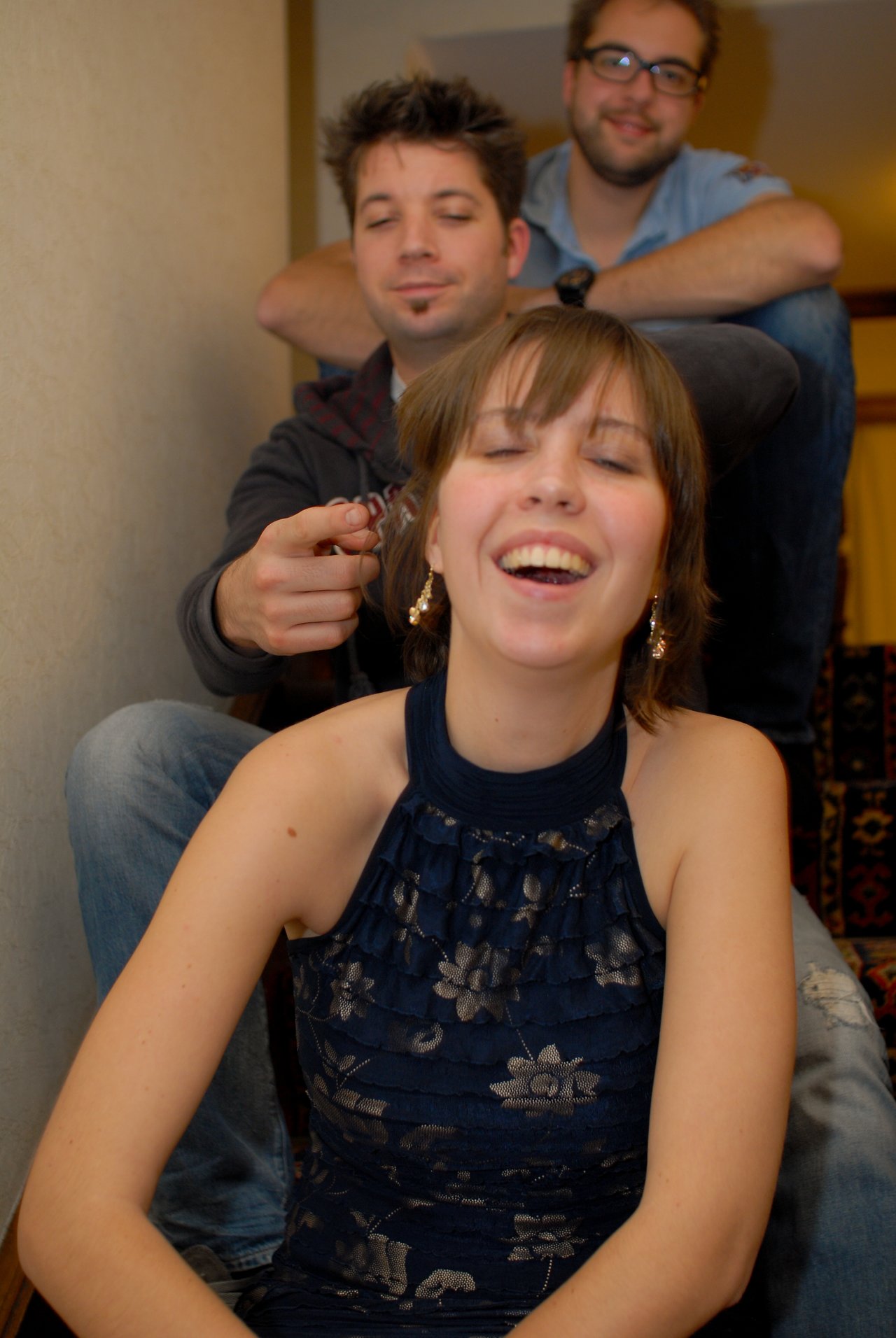 A woman in a blue dress laughs while a man behind her playfully holds a small object near her hair.