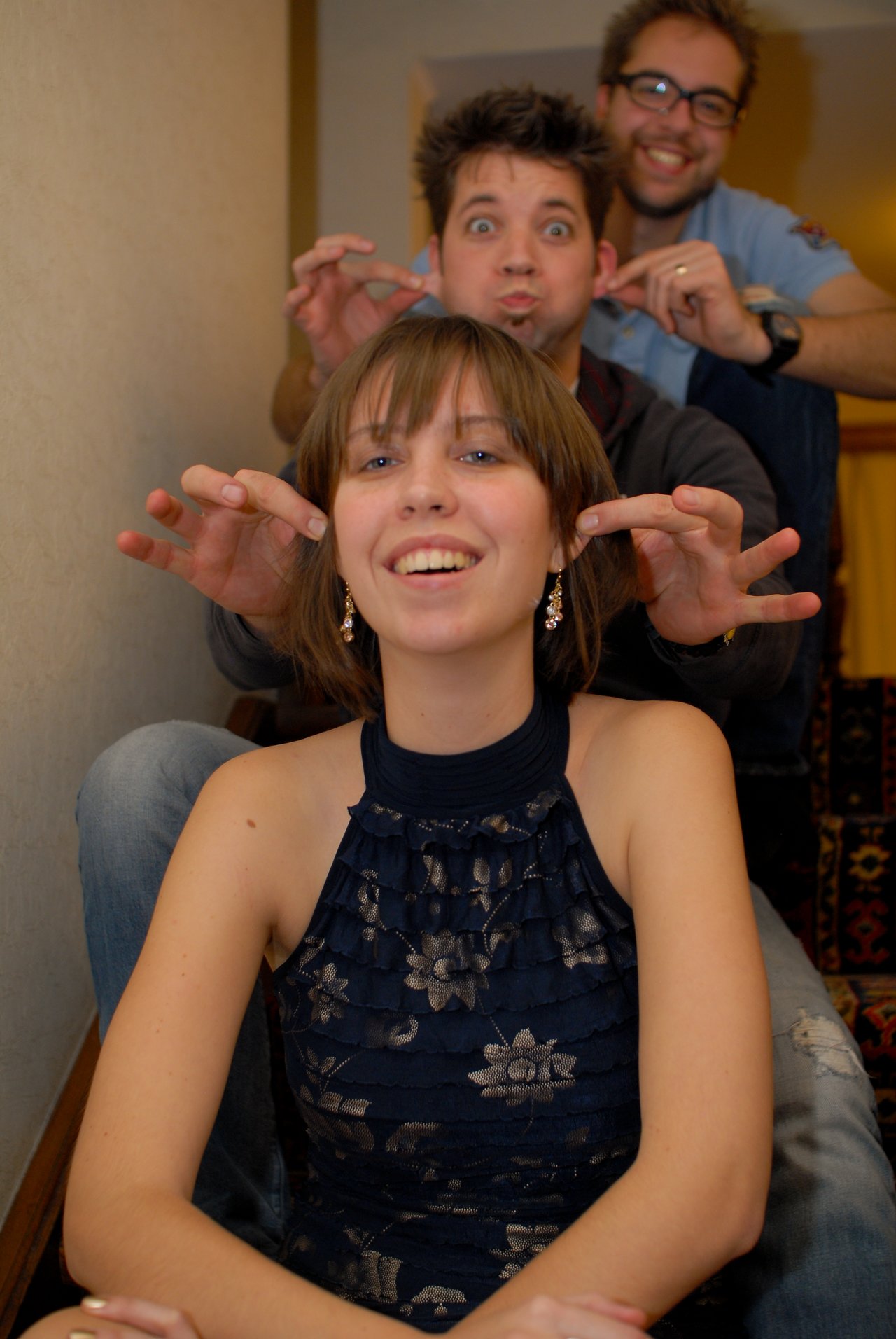 Three people sitting on stairs, playfully making gestures behind the woman in front, who is smiling.
