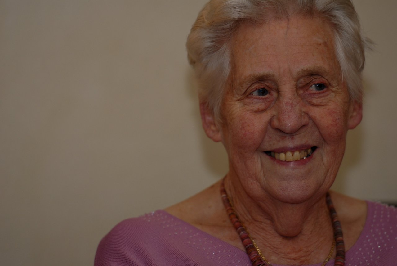An elderly woman with short white hair smiles while wearing a pink top and a beaded necklace.