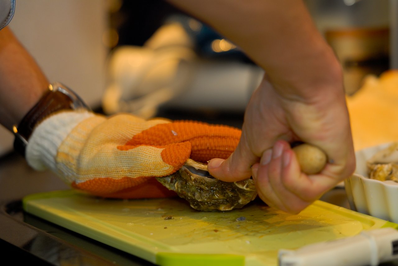 A person wearing protective gloves shucks an oyster using a knife on a cutting board.