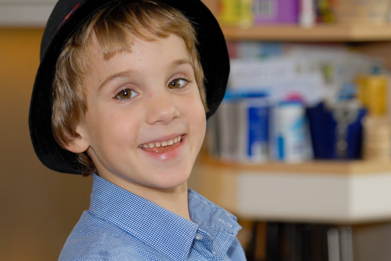 A smiling child wearing a black hat and blue shirt looks at the camera indoors.
