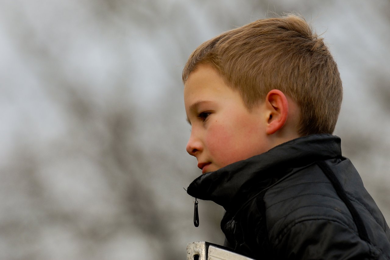 A young boy in a black jacket looks to the side with a neutral expression outdoors.