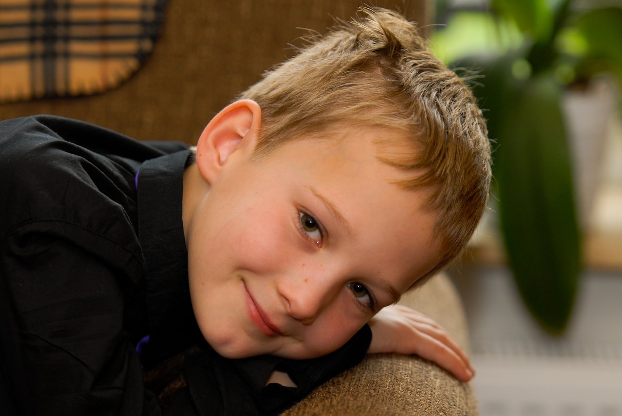 A young child with short blond hair leans on a couch, smiling gently while looking at the camera.