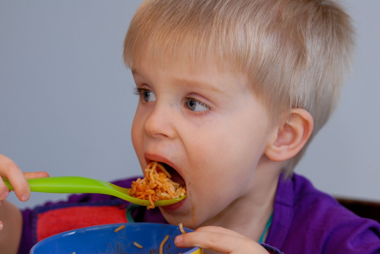 A young child in a purple shirt eats spaghetti with a green spoon from a blue bowl.