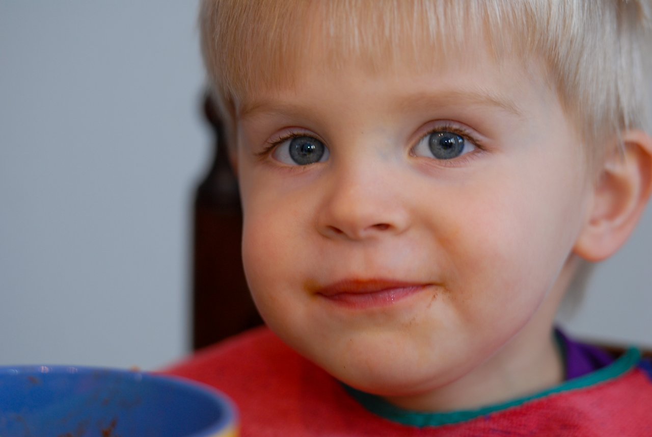 A young child with a messy face sits at the table after eating spaghetti, wearing a red bib.