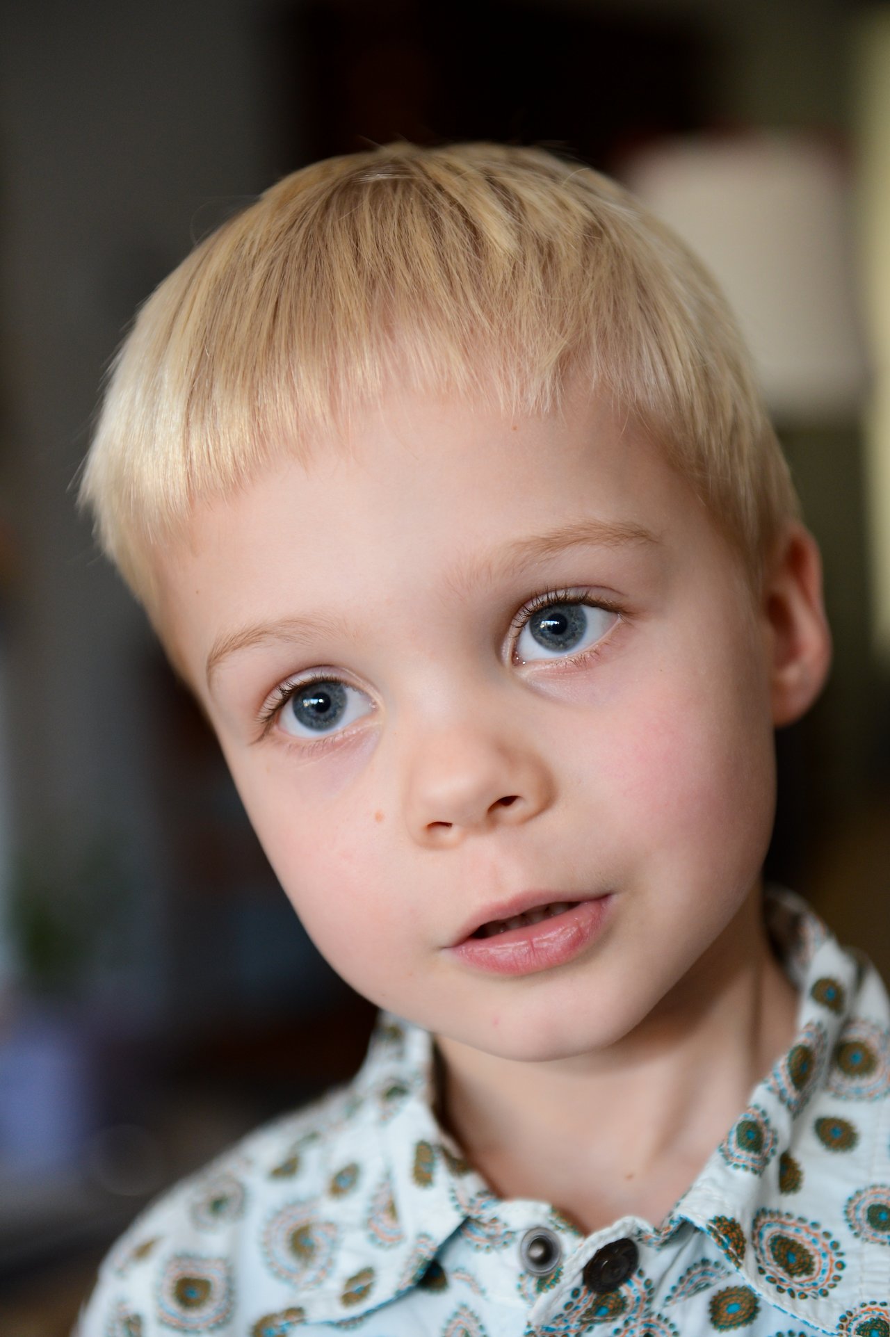 A young child with blonde hair and blue eyes looks slightly to the side, wearing a patterned button-up shirt.