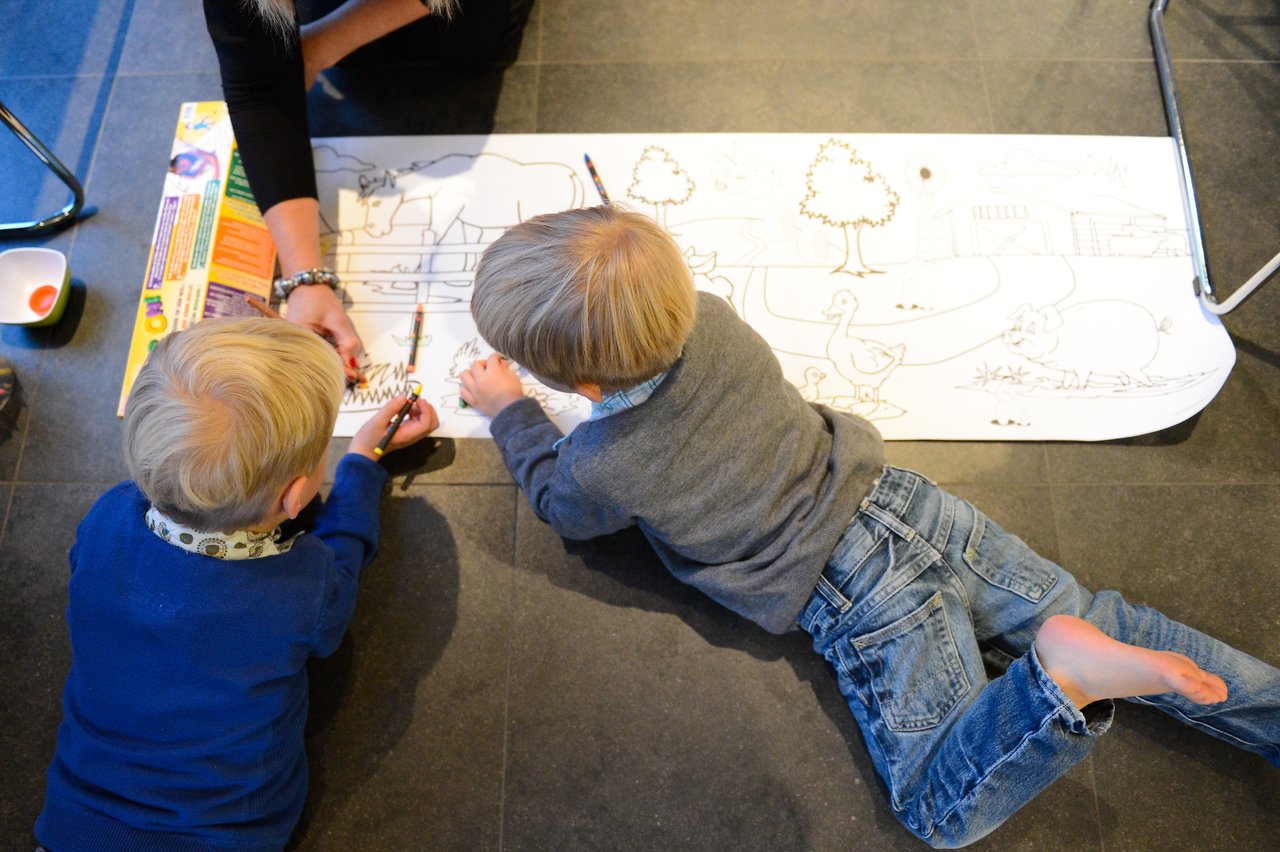 Two children lie on the floor, coloring a large drawing with pencils.