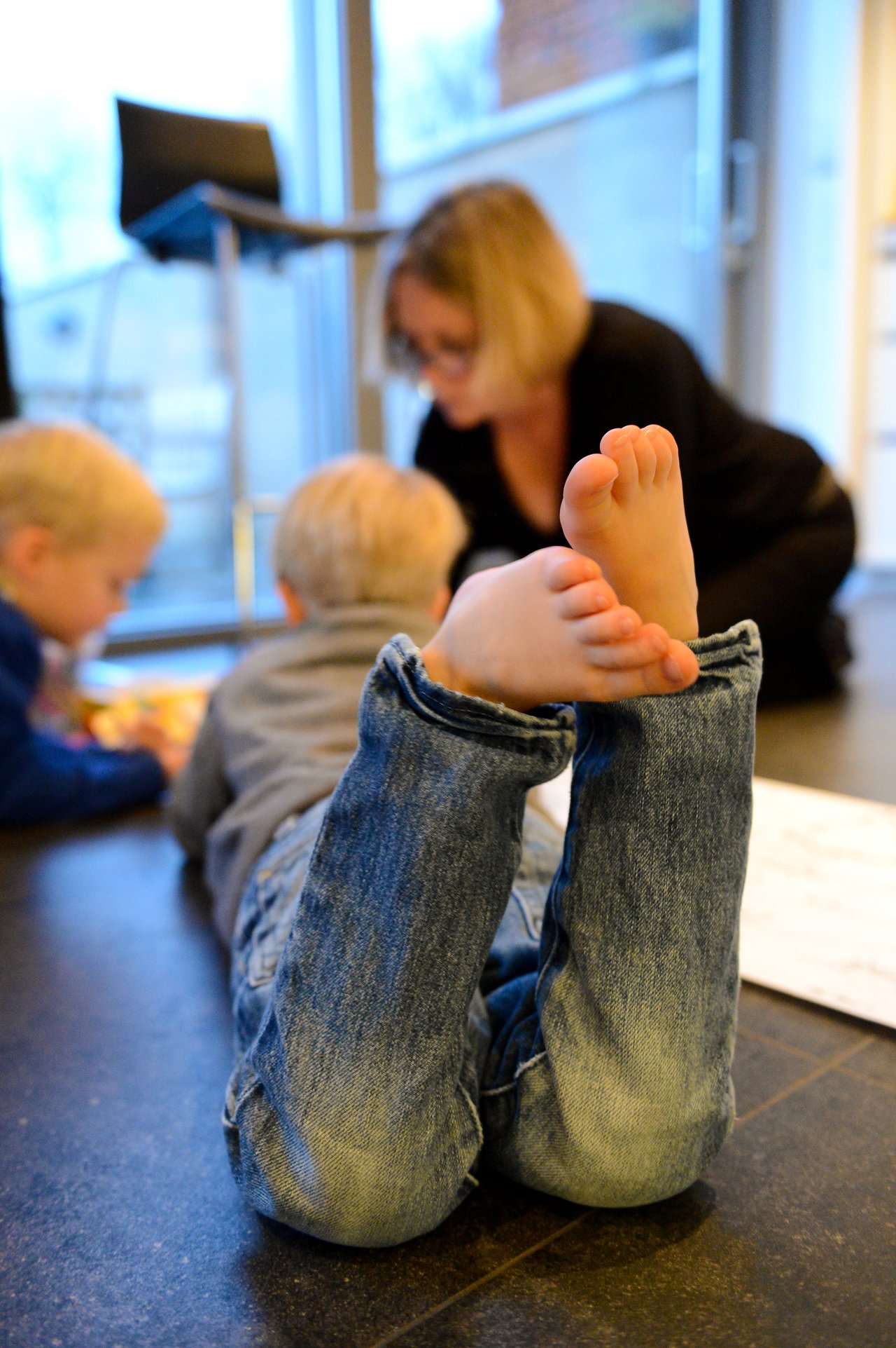 A child lies on the floor barefoot, drawing on paper, with others nearby engaged in the activity.