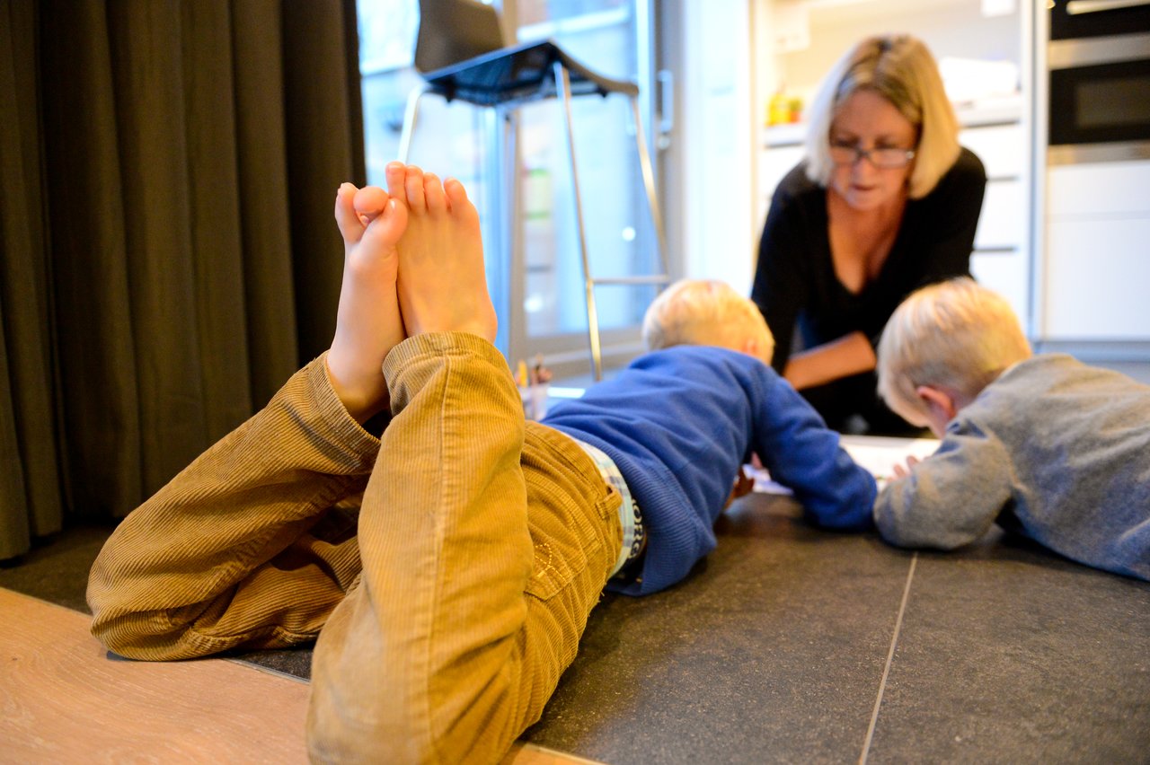 Two children lie on the floor drawing, with bare feet raised, while an adult watches and helps.