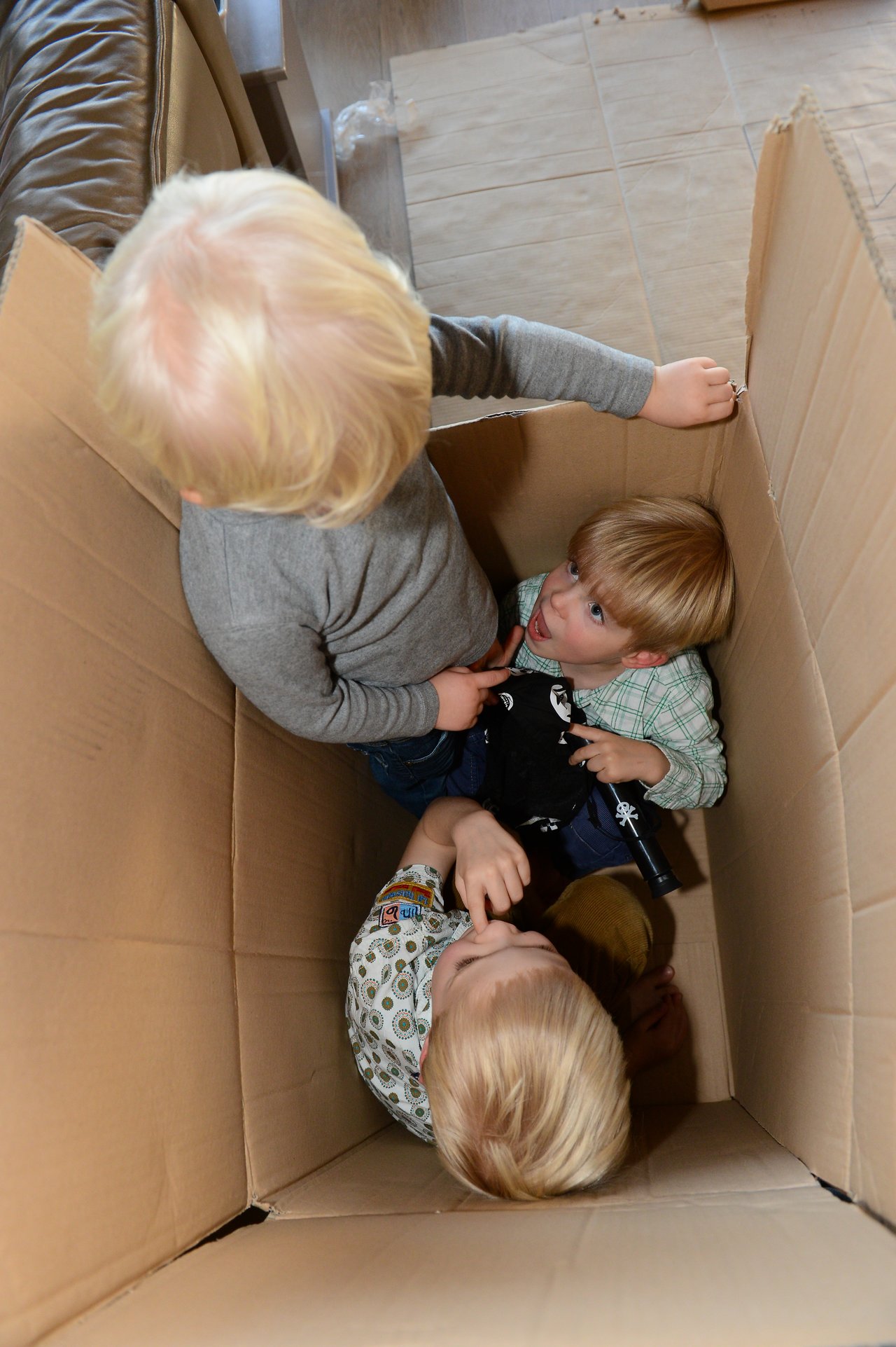 Three young children play inside a large cardboard box, with one standing and two sitting while holding toys.