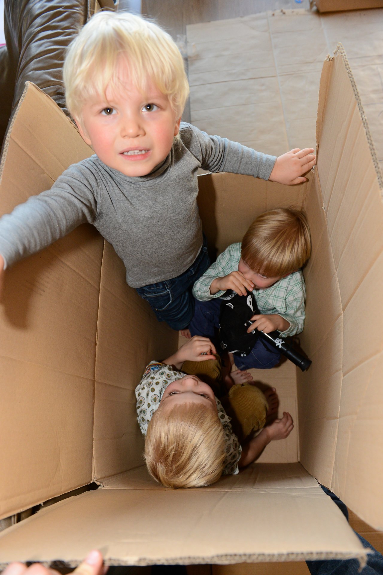 Three young children play inside a large cardboard box, with one standing and two sitting while holding toys.