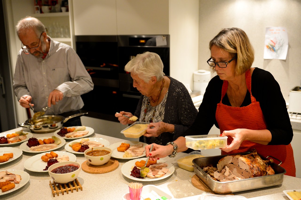 Three people prepare and serve food in a kitchen, plating roasted meat, vegetables, and sauces for a meal.