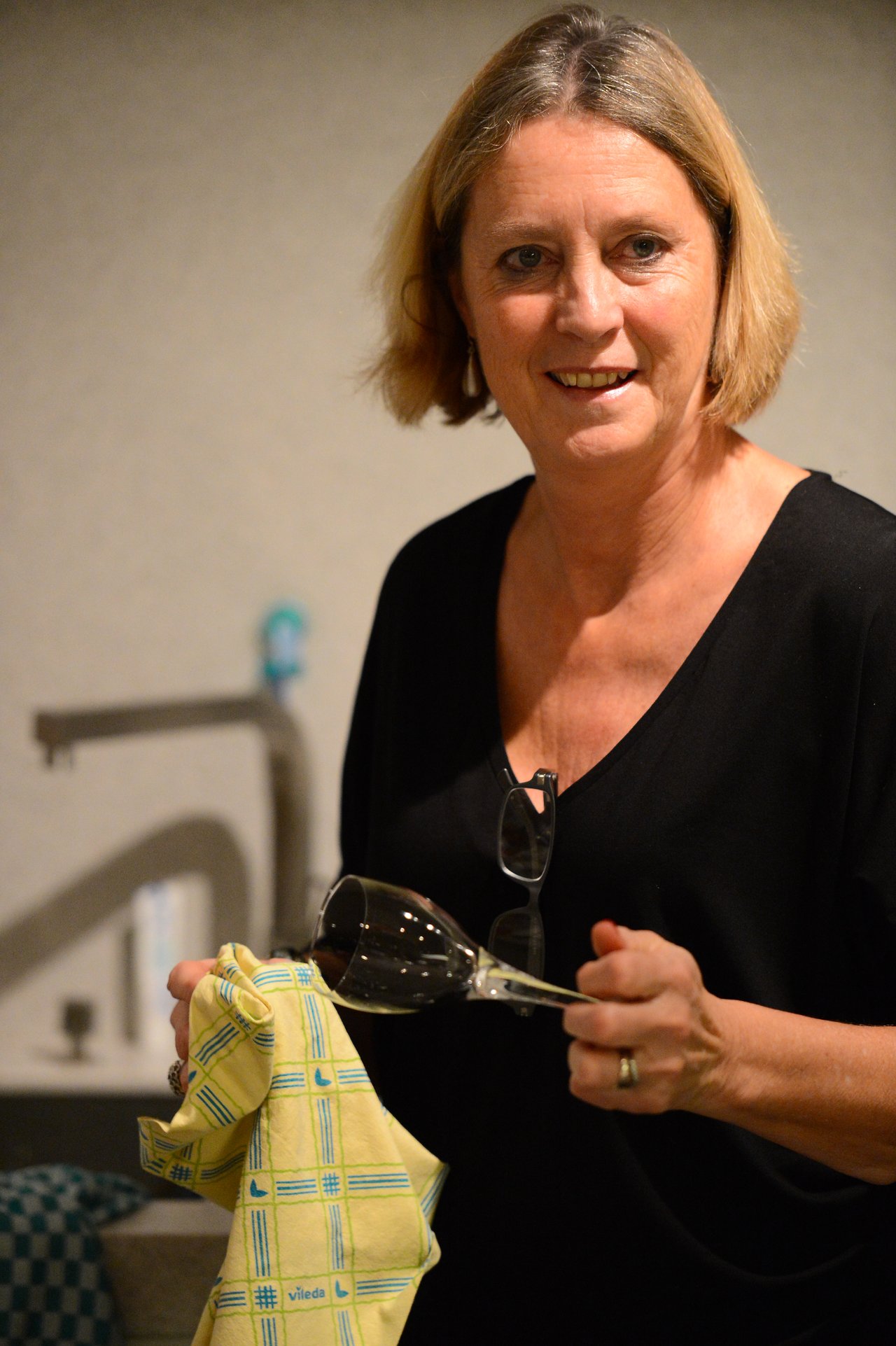A woman in a black shirt dries a wine glass with a yellow cloth in a kitchen.