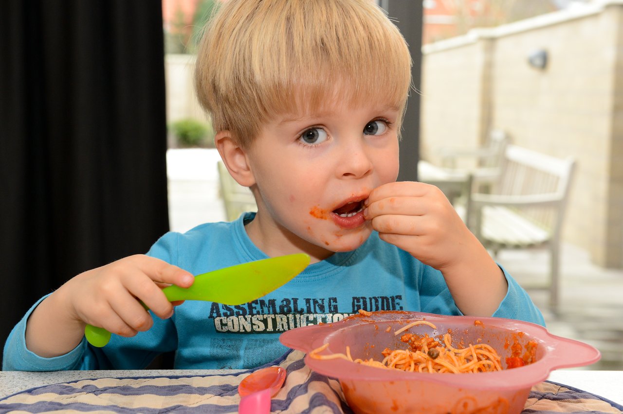 A young child eats spaghetti with their hands, holding a green spoon, with sauce on their face.