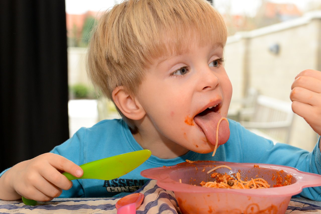 A young child in a blue shirt eats spaghetti, playfully sticking out their tongue with a noodle on it.