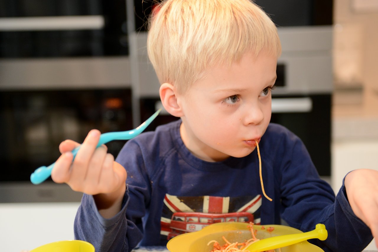 A young child in a blue shirt eats spaghetti from a yellow bowl, holding a fork in one hand.