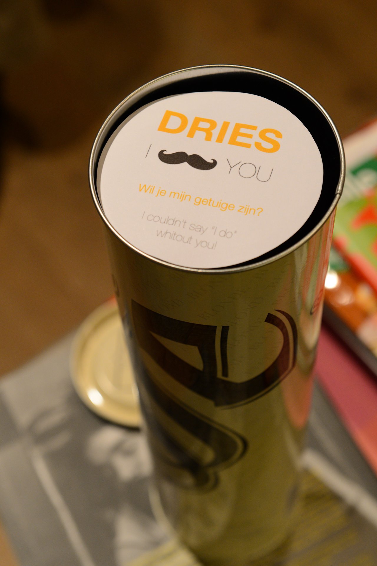A cylindrical gift container with a message asking "Dries" to be a wedding witness, featuring a mustache graphic.