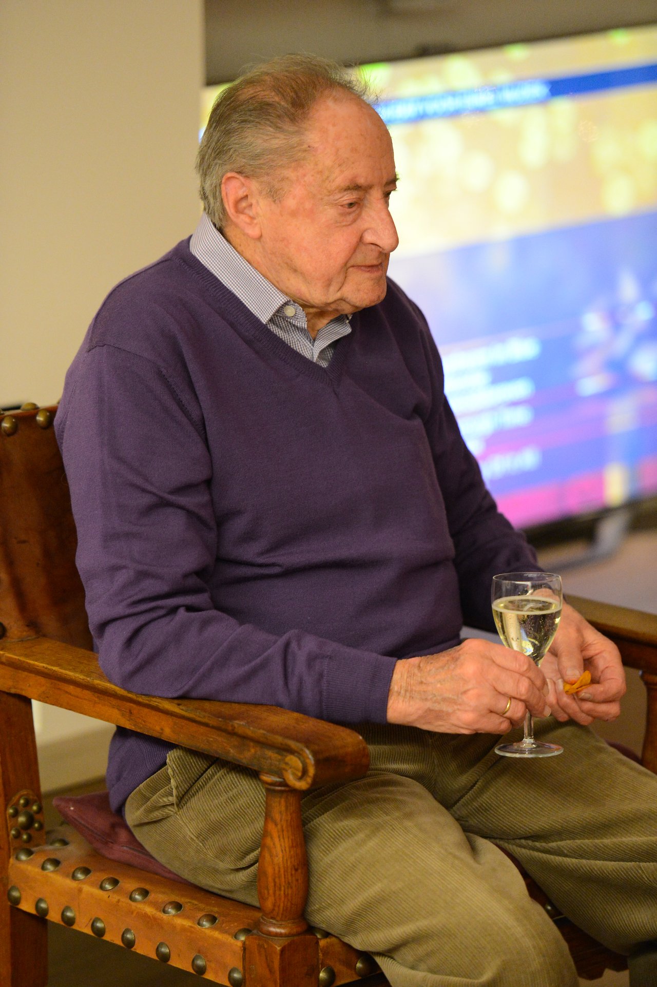 Elderly man sitting in a wooden chair, holding a glass of white wine and a small snack.