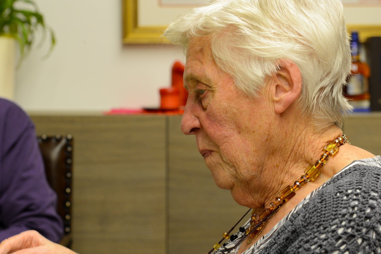 Elderly woman with short white hair wearing a beaded necklace, looking down in a thoughtful expression.