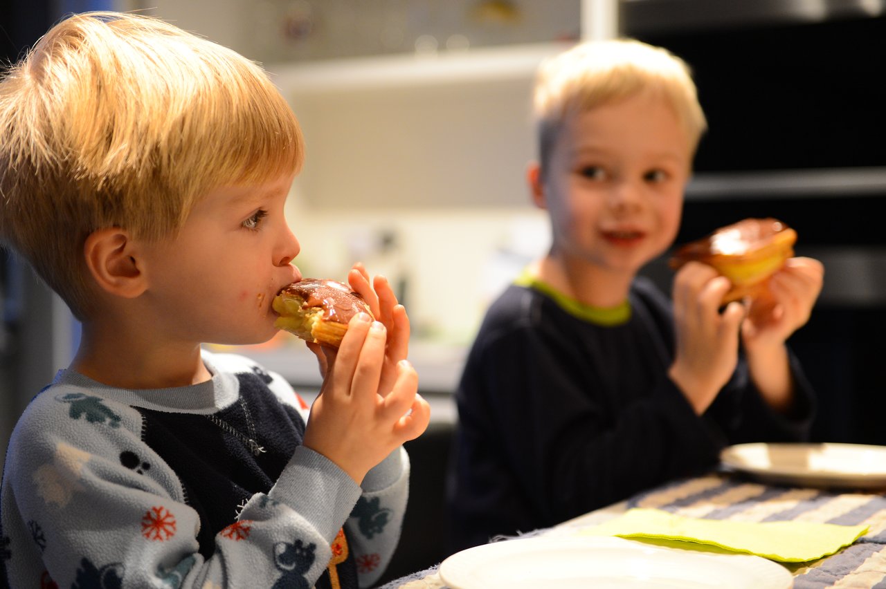 Two young children in pajamas eat chocolate-covered pastries at a table, one taking a bite while the other smiles.
