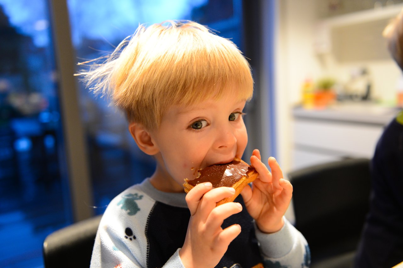 A young child in pajamas takes a bite of a chocolate-covered pastry at the table.
