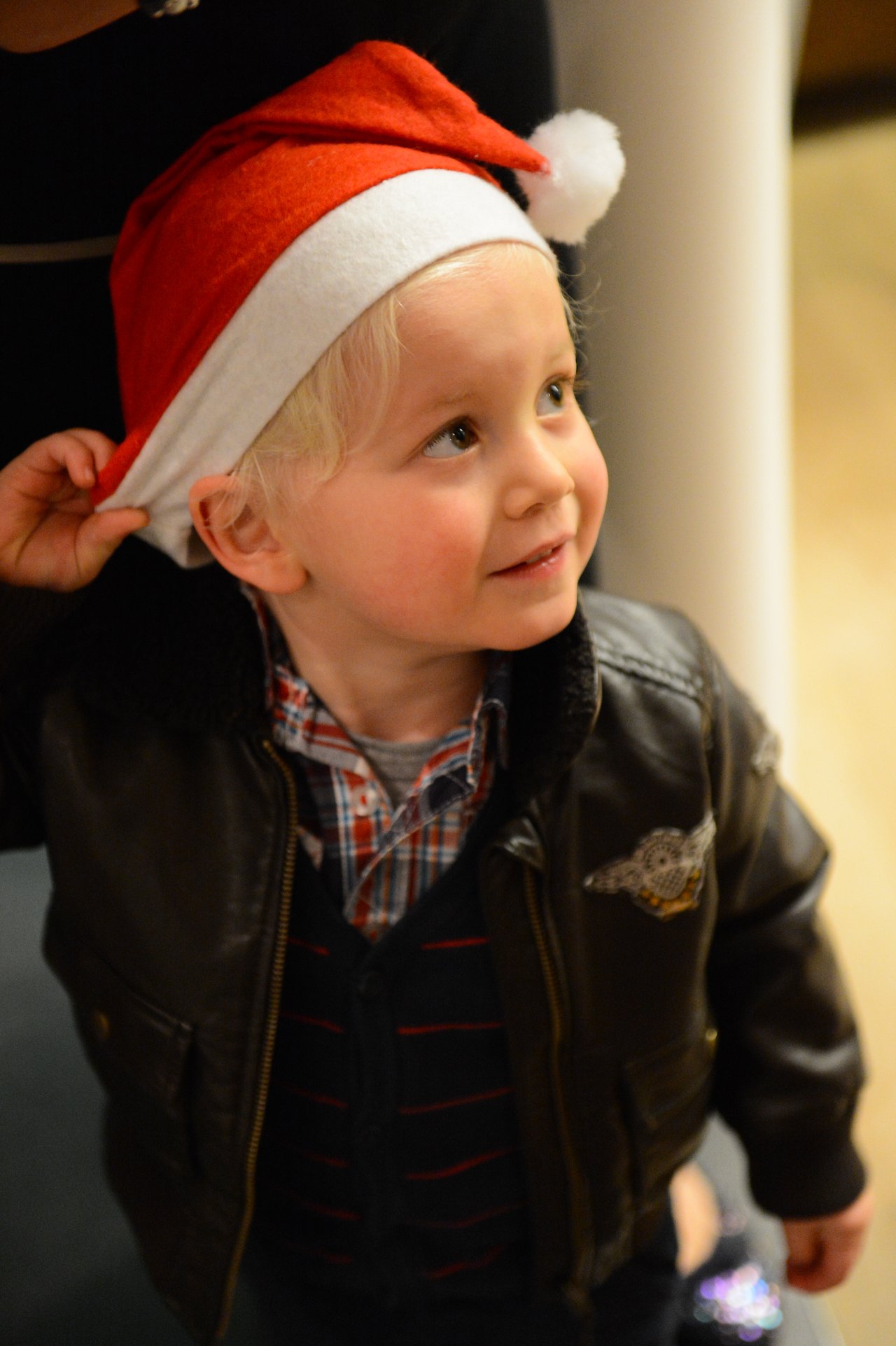 A young child wearing a Santa hat and leather jacket looks up while adjusting the hat with one hand.