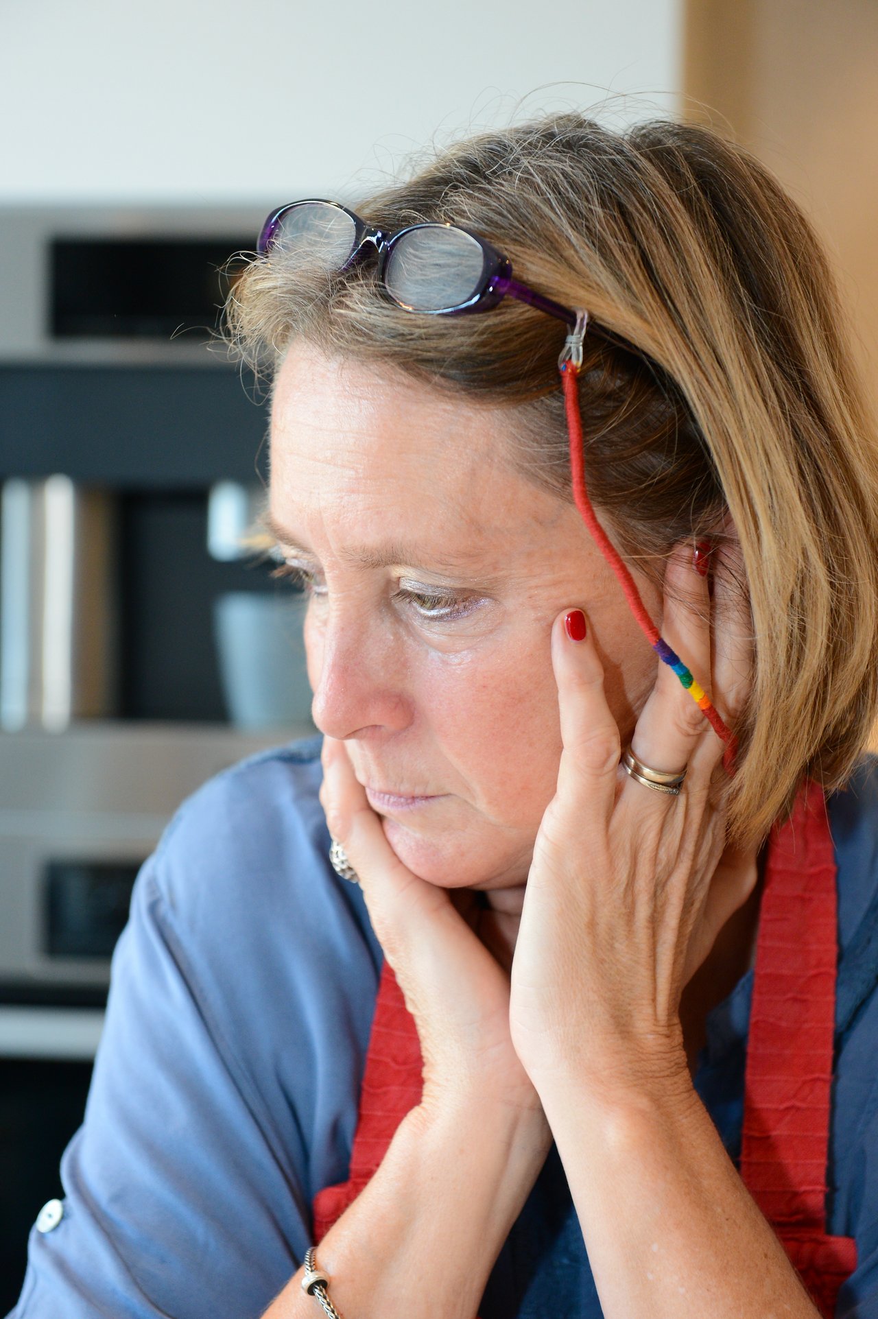 A woman wearing glasses on her head and a red apron rests her hands on her face, looking thoughtful.