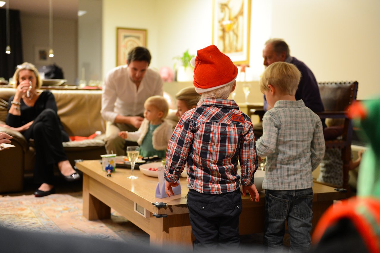 Children in festive outfits stand near a table, while adults sit and chat in a warmly lit living room.