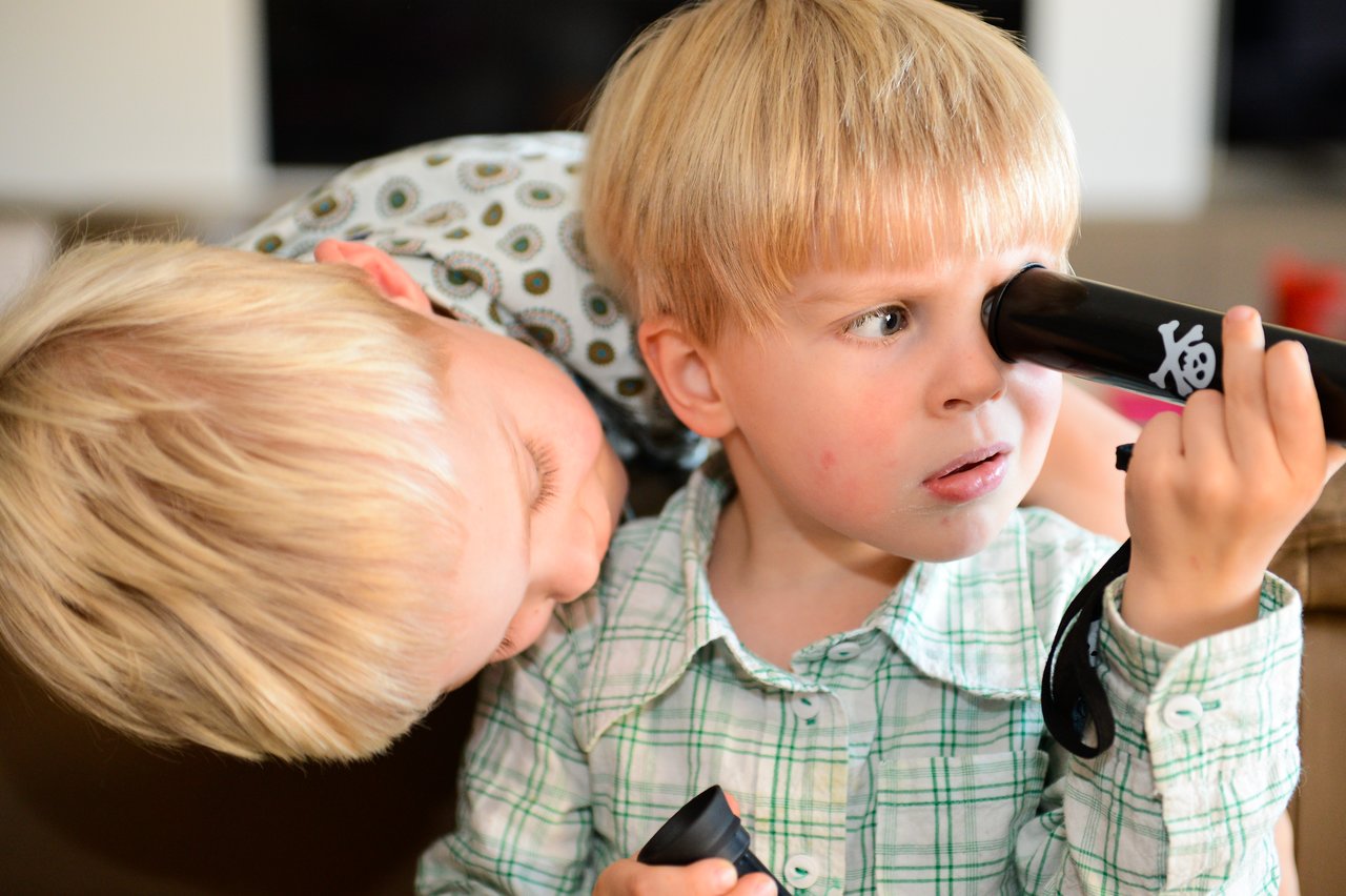 A young boy looks through a toy pirate telescope while another child leans in closely beside him.