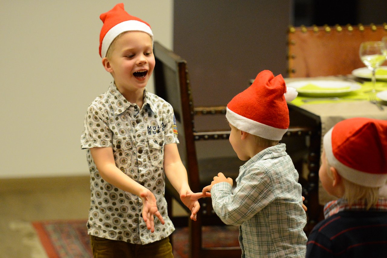 Three young children wearing Santa hats are playing together indoors, smiling and laughing.