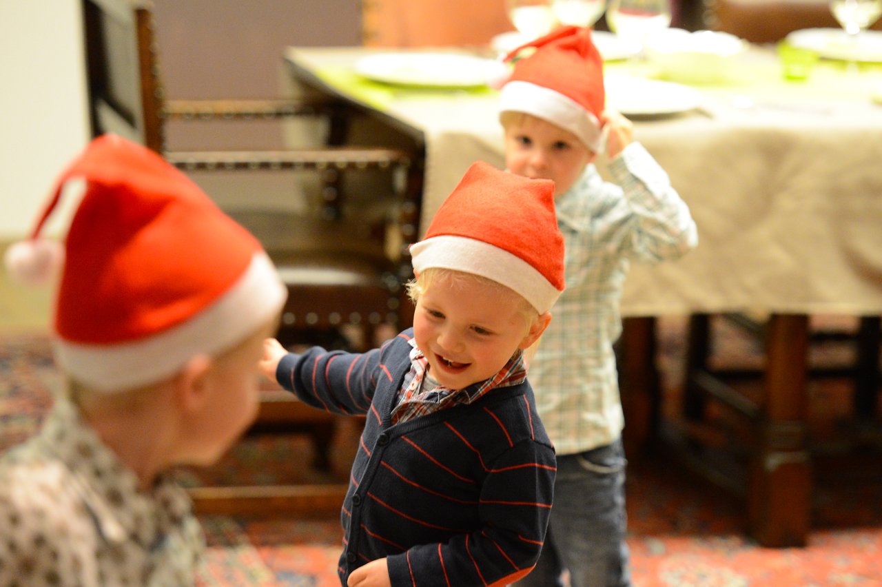 Three young children wearing Santa hats are playing indoors, smiling and interacting with each other in a festive setting.