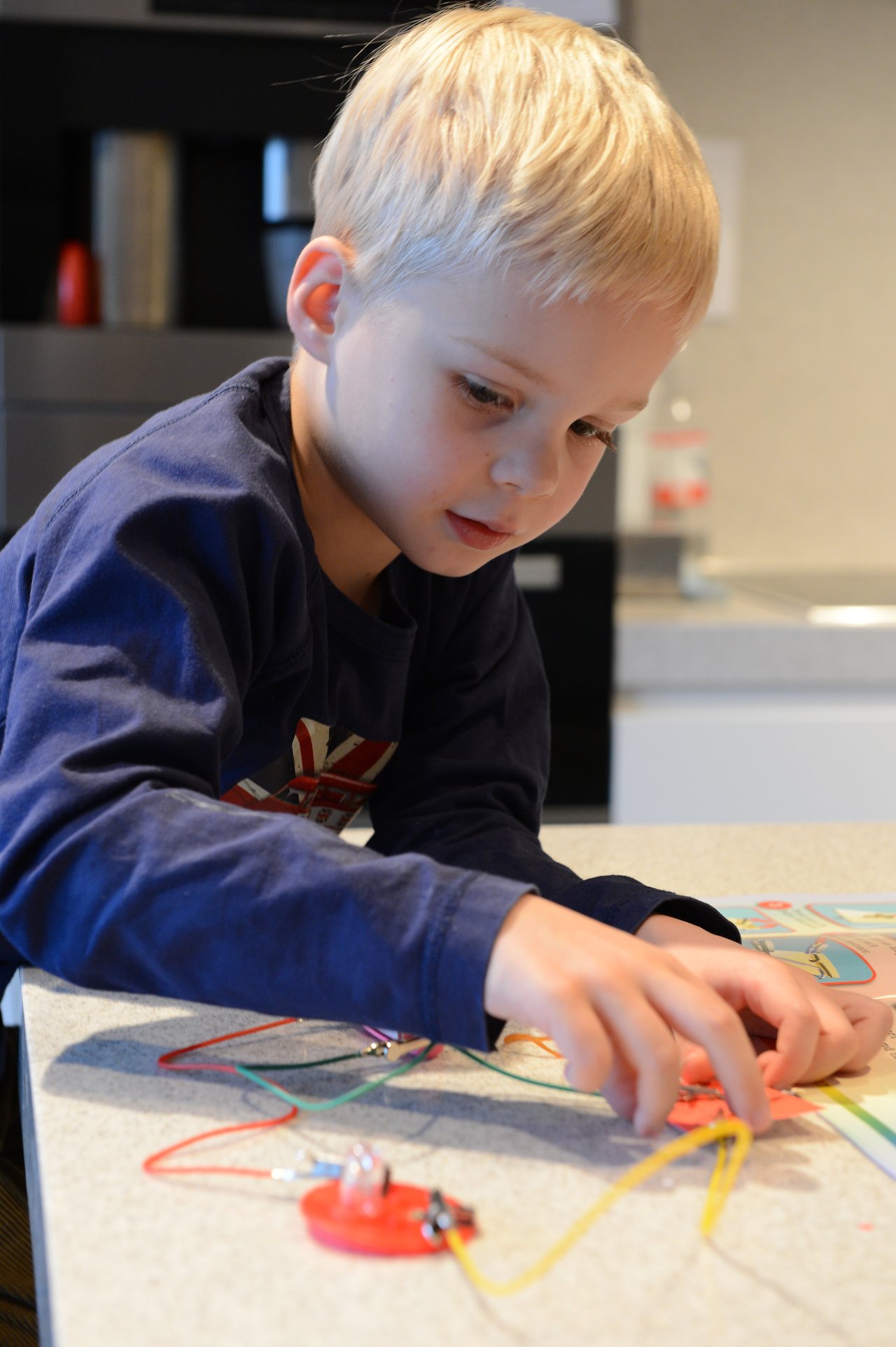 A young child connects wires to a small circuit board, focusing on an electrical experiment at a table.